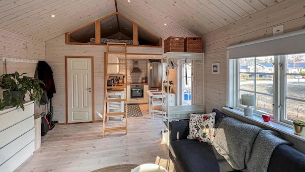 A cozy cabin interior with a loft bedroom, a kitchen, and a living area with a couch and large windows.
