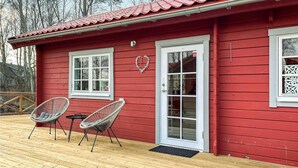 A red wooden cabin with two windows and a door, a porch with two wicker chairs, and a wooden deck.