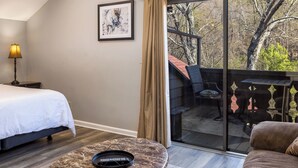 A hotel room with a bed, a desk with a lamp, a marble table, a chair, and a balcony with a view of trees.
