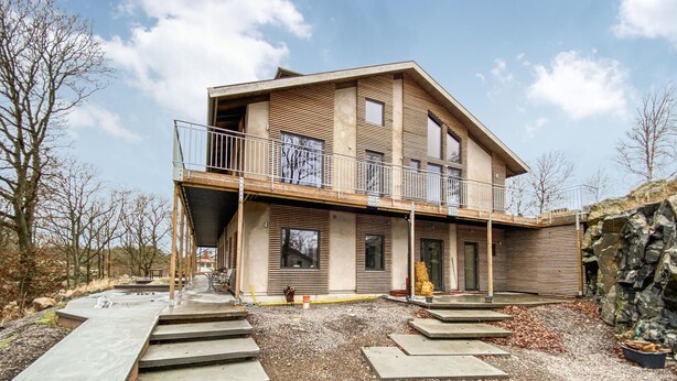 Une maison moderne à deux étages, dotée d’un balcon, d’un mur en pierre et d’une allée gravillonnée.