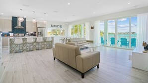 A modern living room with a beige sofa, a glass coffee table, and a kitchen area with a bar. - Oceanfront PARADISE w Heated pool, 55’ dock, ocean view & bathroom in every room (Marathon)