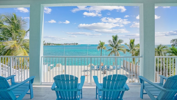 A balcony with blue Adirondack chairs facing a beach with clear turquoise water and palm trees. - Oceanfront PARADISE w Heated pool, 55’ dock, ocean view & bathroom in every room (Marathon)
