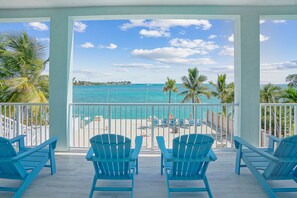 A balcony with blue Adirondack chairs facing a beach with clear turquoise water and palm trees. - Oceanfront PARADISE w Heated pool, 55’ dock, ocean view & bathroom in every room (Marathon)