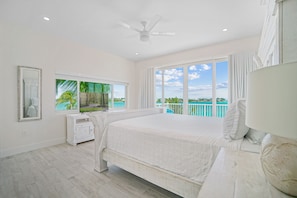 A bedroom with a large bed, a ceiling fan, a window with a view of the ocean, and a white dresser.