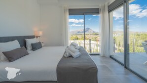 Une chambre d’hôtel moderne avec un grand lit, un balcon offrant une vue sur les montagnes et un panorama de la ville en arrière-plan.