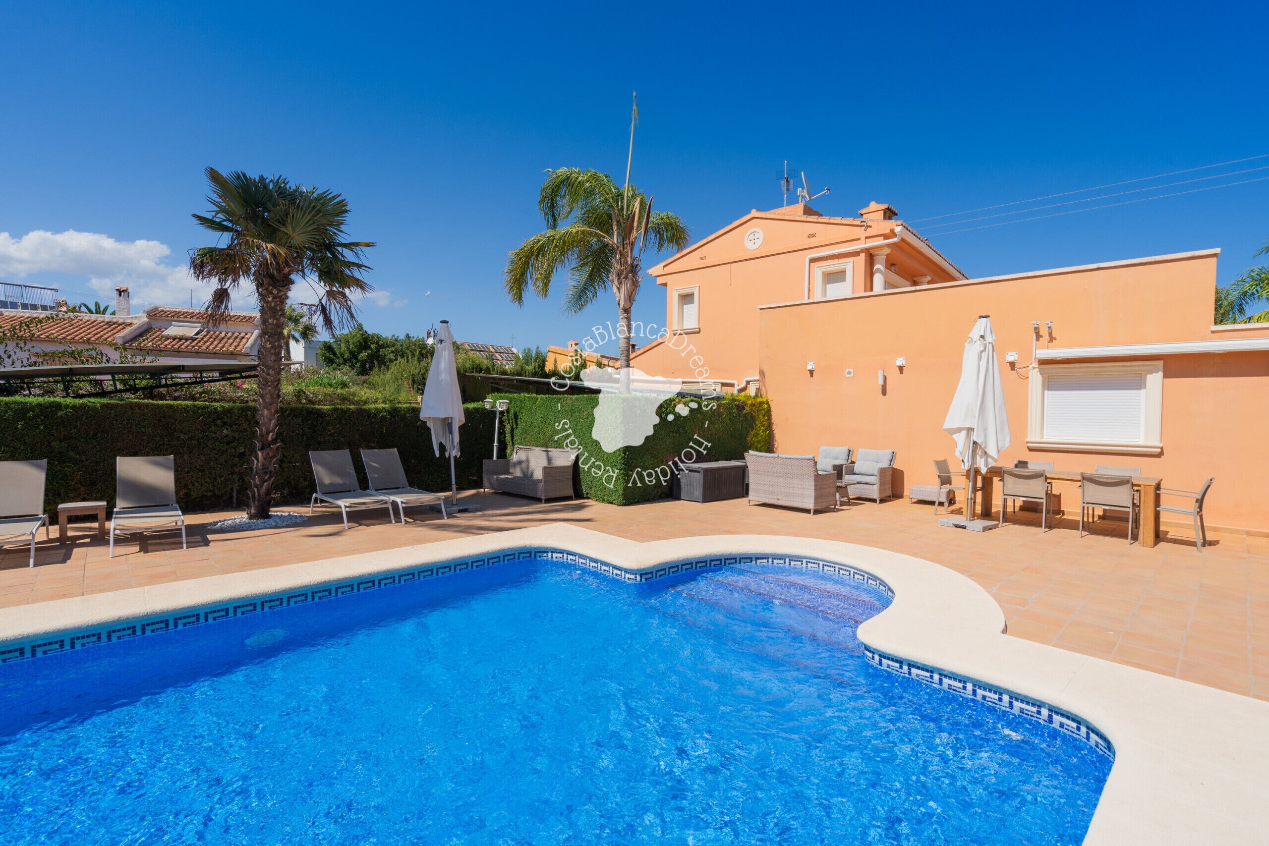 A swimming pool with lounge chairs and umbrellas, a building with a tiled roof, and a clear blue sky.