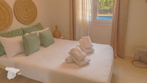 Een netjes opgemaakt bed met witte lakens en opgerolde handdoeken, een raam met gordijnen en uitzicht op groen buiten.