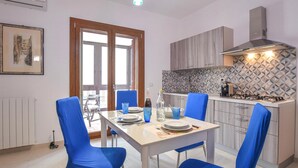 A compact kitchen with a dining area, featuring a table set for two with blue chairs, a gas stove, and a patterned backsplash. - 1 bedroom beautiful apartment in Siracusa (Siracusa)