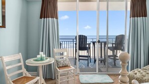 Ein Balkon mit Blick auf den Ozean, ausgestattet mit Stühlen und einem Tisch, und eine Glastür zum Strand.