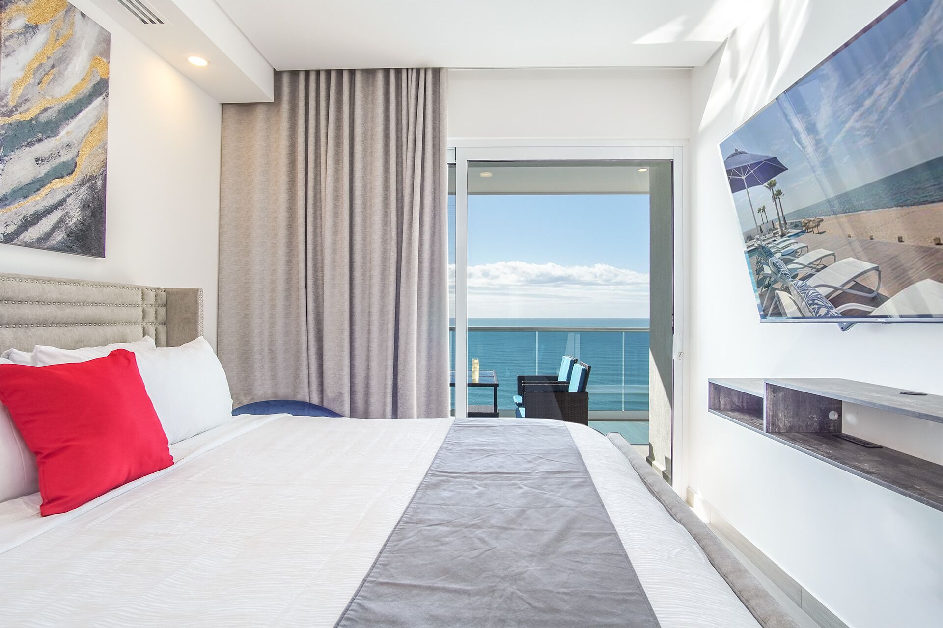 A hotel room with a bed, a view of the sea, a painting, and a window with curtains.