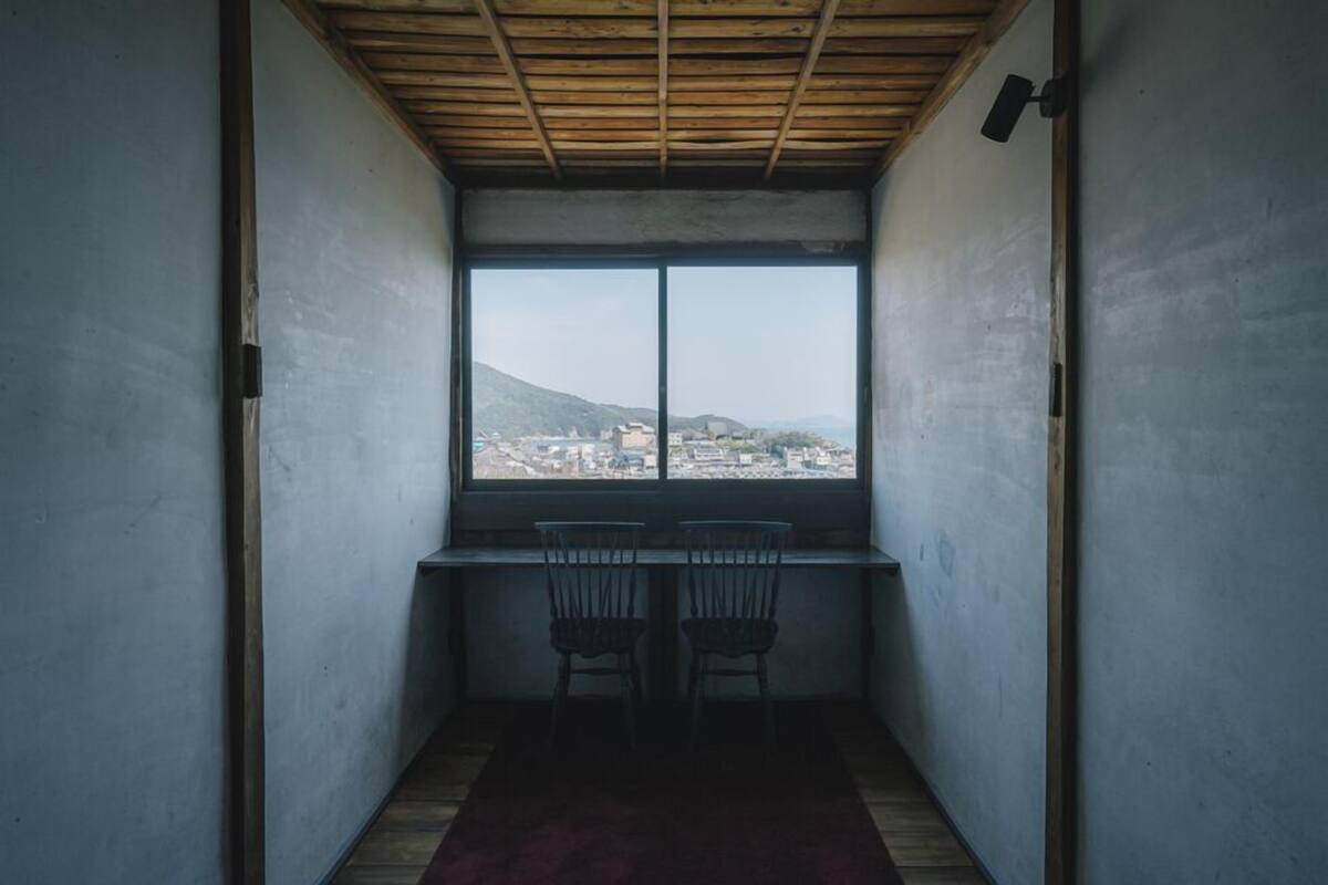 Quarto com uma janela grande, duas cadeiras, uma escrivaninha e teto de madeira.