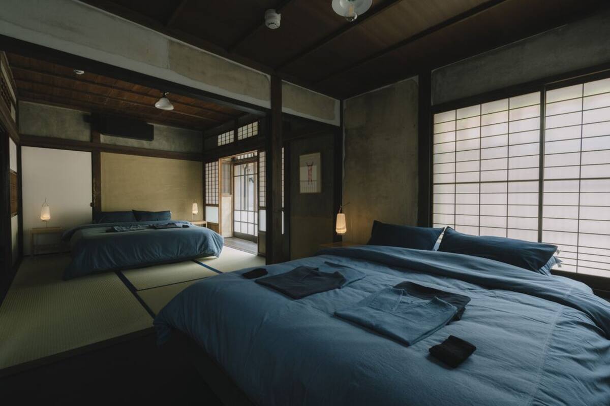 Quarto tradicional japonês com tatames, uma cama grande com roupa de cama azul, uma pequena mesa e uma porta de correr.