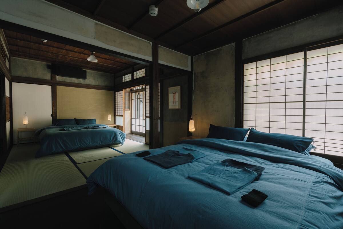 Um quarto em estilo japonês tradicional, com tatames, duas camas com roupa de cama azul e uma porta de papel shoji.