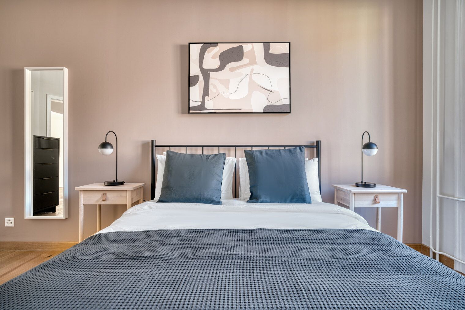 A bedroom with a bed, two bedside tables, a mirror, and a wall-mounted abstract artwork.