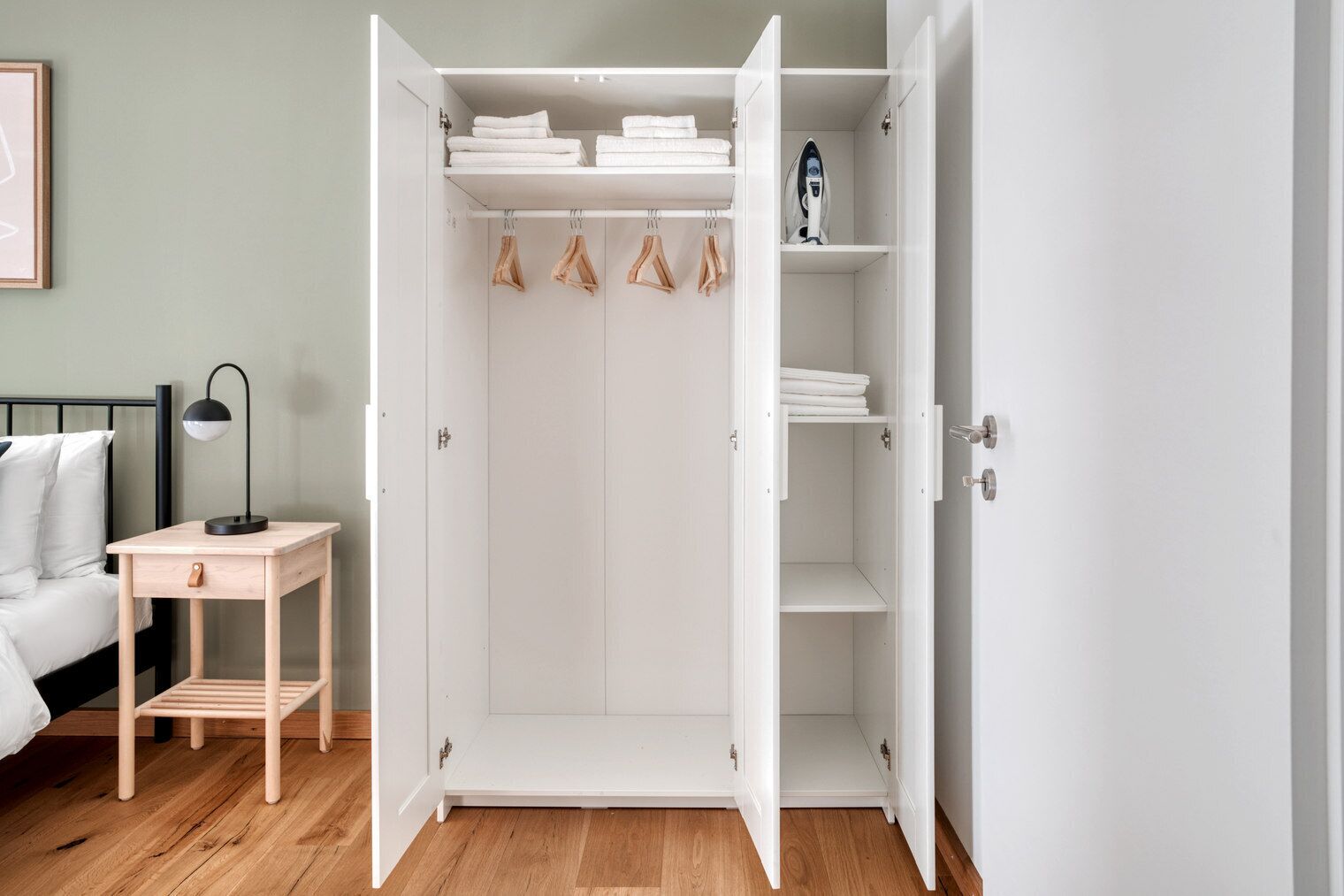 Un dormitorio con un armario blanco, una mesita de noche con lámpara y una cama con ropa de cama blanca.
