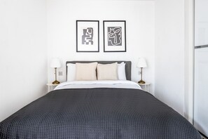 A neatly made bed with a dark grey bedspread and white pillows, flanked by two bedside tables with lamps, in a room with white walls and two framed abstract artworks above the bed.