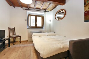 A bedroom with a bed, a chair, a TV, a window, and a ceiling with exposed beams.