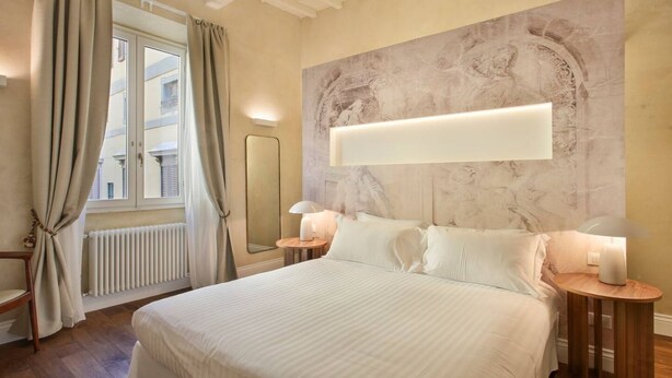 Ein Hotelzimmer mit einem großen Bett, einem Marmorkopfschild, einem Nachttisch mit Lampe, einem Fenster mit Vorhängen und einem Heizkörper.