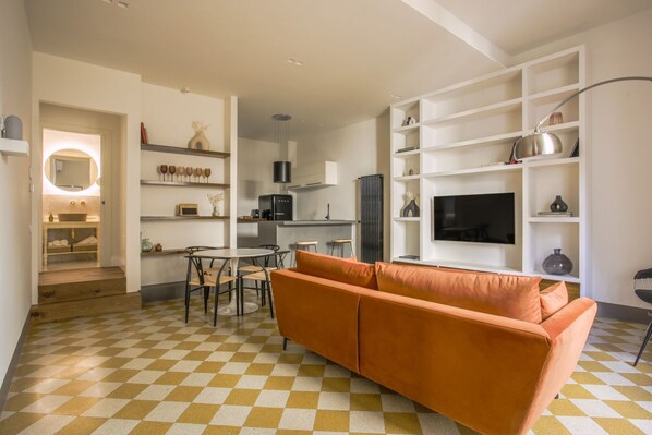 A modern living room with a checkered floor, a sofa, a dining table with chairs, a kitchen area, and a TV on a white shelf.