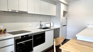 A modern kitchen with white cabinetry, a built-in oven, a dishwasher, and a wooden dining table. - Sixty Six Boutique Apartments (North Hobart)