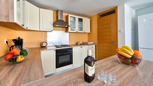Une petite cuisine équipée d’une cuisinière, d’un four et d’une machine à café. On peut voir une bouteille de whisky Ballantine’s et un saladier de fruits sur le comptoir.