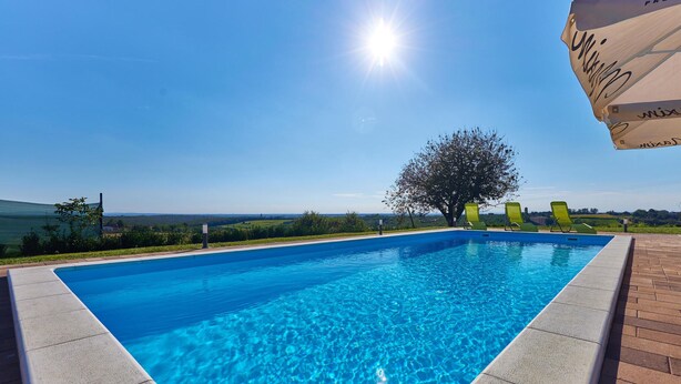 Une piscine rectangulaire à l’eau d’un bleu limpide, entourée d’une terrasse en briques. Trois chaises longues vertes sont alignées le long du bord de la piscine. Un grand parasol est partiellement visible dans le coin supérieur droit.