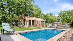 A rectangular swimming pool with a beige deck, surrounded by a grassy yard and trees. - Hein House | Hot Tub | Shared Pool (Fredericksburg)