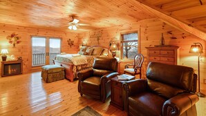 A cozy log cabin interior with a bed, leather chairs, a fireplace, and a ceiling fan. - Family Cabin With Mountain Views (Boone)