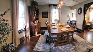 Comedor con mesa de madera dispuesta para cuatro personas, sillas a los lados, un mueble de madera oscura y un ventanal con cortinas.