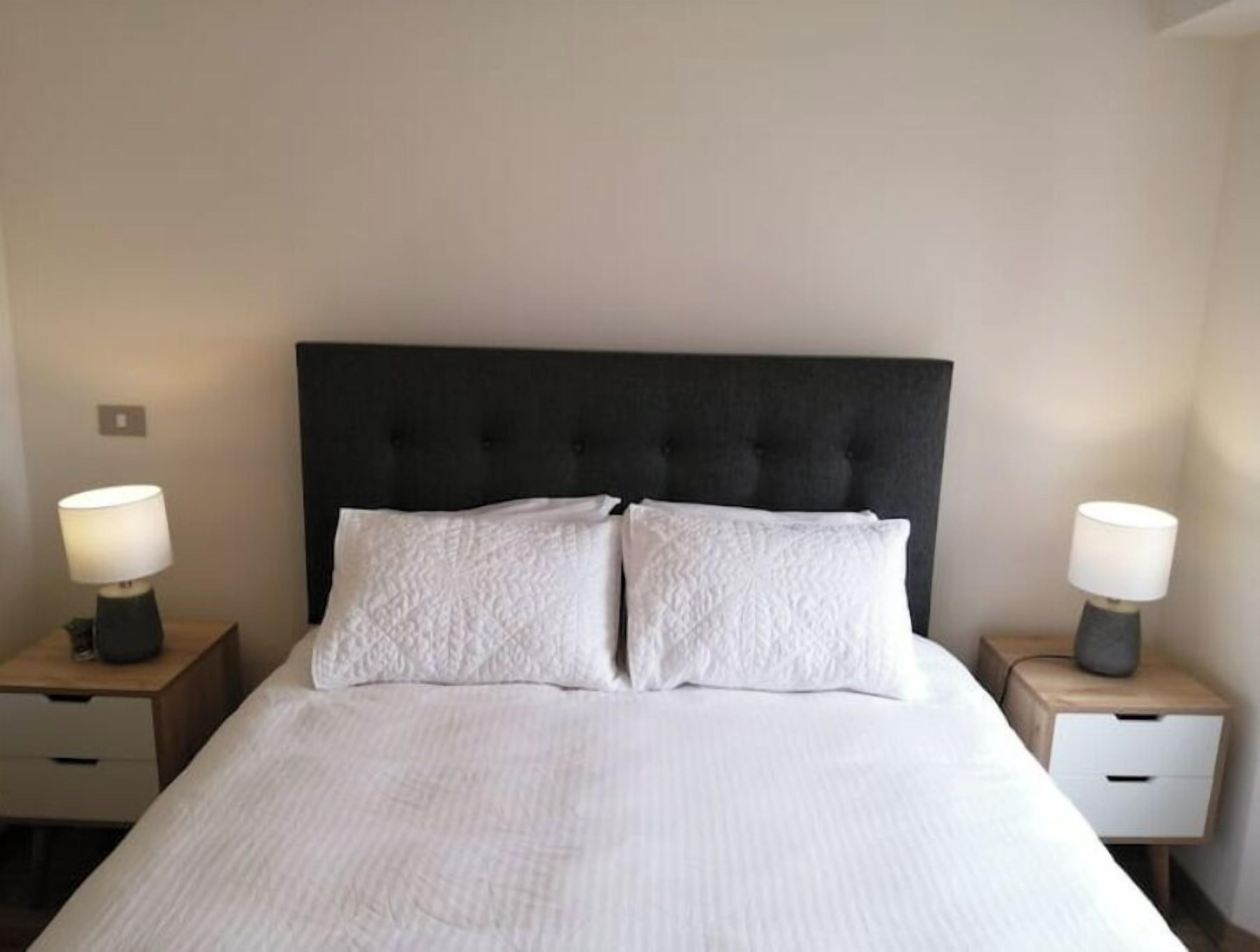 A neatly made bed with a dark headboard, two bedside tables with lamps, and a plain wall.