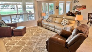 A spacious living room with a large window, leather sofas, a patterned rug, and a dining area visible through the glass doors. - PC Furn Golf Course Home. 36 PB courts for Nat's Nov `24 incl cart, hk 'ing util (Goodyear)