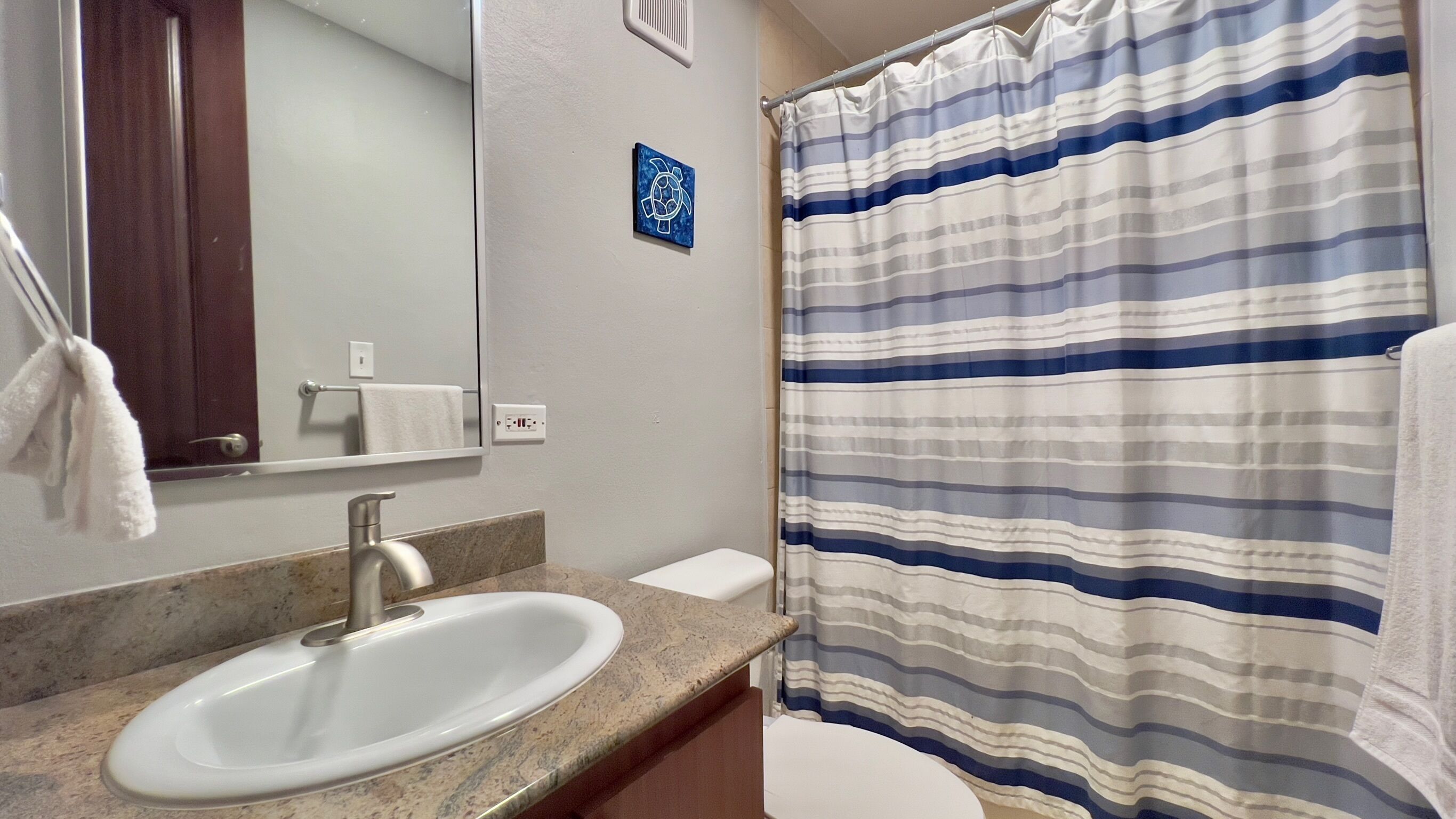 A bathroom with a white sink, a mirror, a towel rack, a striped shower curtain, and a small blue artwork on the wall.