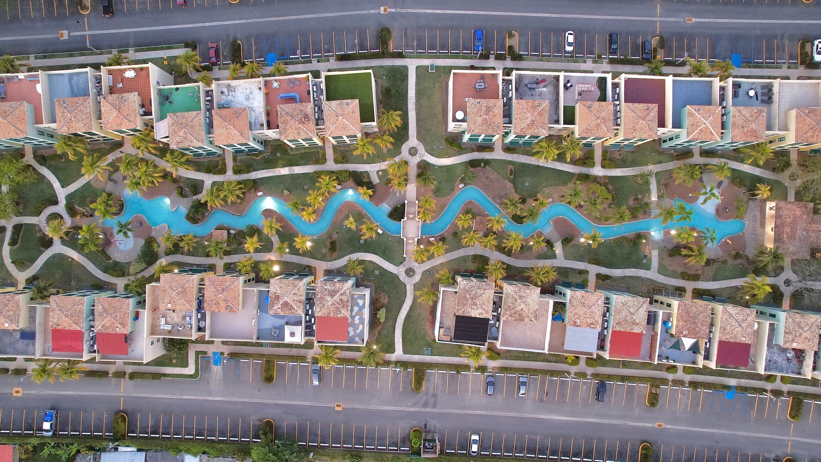 Vue aérienne d’un complexe hôtelier comprenant plusieurs bâtiments, une piscine centrale et un parking.