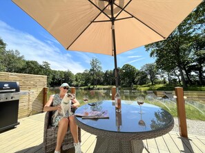 A person sitting at a round glass table with a large beige umbrella, a grill, and a glass of wine on a wooden deck overlooking a lake. - Rowan Boutique Lodge for couples with a dog (York)