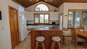 A kitchen with white cabinets, a wooden island with stools, a large window, and a dining area with chairs. - PLEASURE ISLAND - 5 BR Home in Schooner Ridge w/Access to Community Pools & Rec Center (Duck)