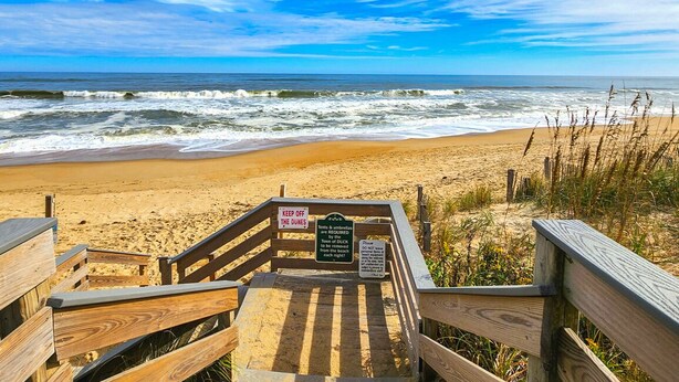 Une promenade en bois mène à une plage où un panneau indique qu’il est nécessaire d’avoir des tentes et des parasols.