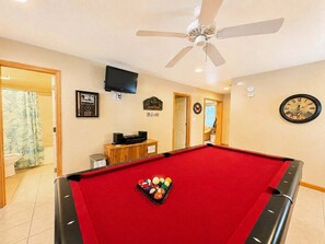 A pool table with balls, a ceiling fan, a clock, and a television in a room with beige walls and a wooden door. - Driftin South - 5 BR Oceanside Home in Duck - Short Walk to Shops & Restaurants (Duck)