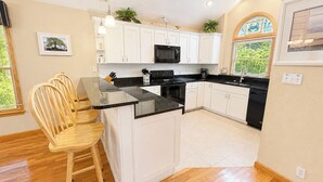 A modern kitchen with white cabinets, a black countertop, and a wooden dining area. - Driftin South - 5 BR Oceanside Home in Duck - Short Walk to Shops & Restaurants (Duck)
