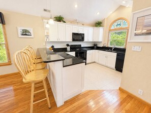 A modern kitchen with white cabinets, a black countertop, and a wooden dining area. - Driftin South - 5 BR Oceanside Home in Duck - Short Walk to Shops & Restaurants (Duck)
