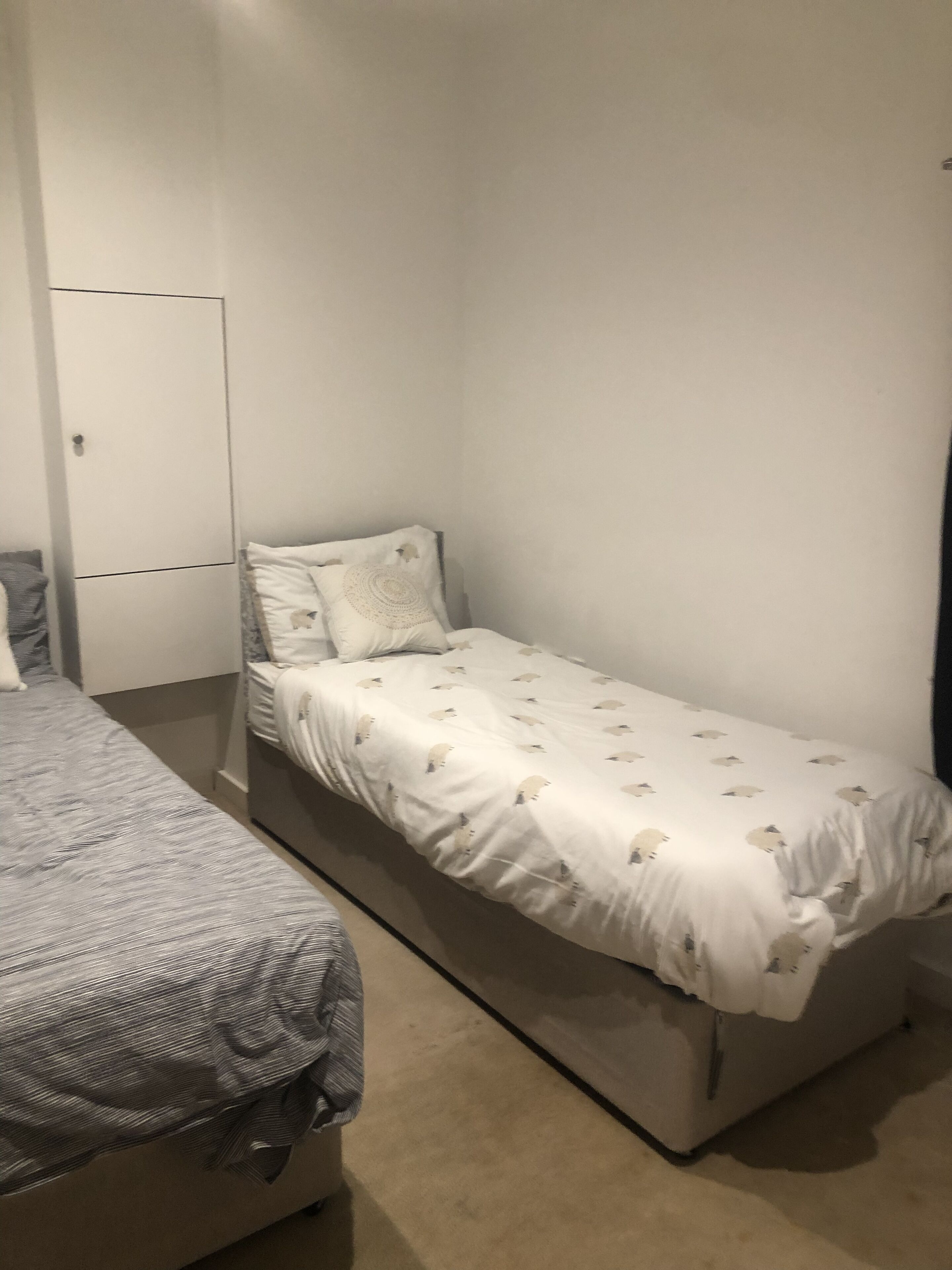 A single-bed room with a plain white wall, a small bedside table, and a grey bedspread.