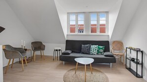 En moderne loftlejlighed med skråvæg, en grå sofa med mønstrede puder, et lille hvidt bord og en fletstol.