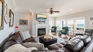 A spacious living room with a fireplace, leather sofas, a television, and a view of the ocean through large windows. - Hobbs Realty - Enjoy "Miss B Haven", a pet-friendly oceanfront home w/ private pool (Holden Beach)