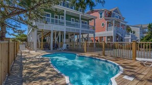 A pool area with a wooden deck, lounge chairs, and a two-story house with a balcony. - Hobbs Realty - Enjoy "Miss B Haven", a pet-friendly oceanfront home w/ private pool (Holden Beach)