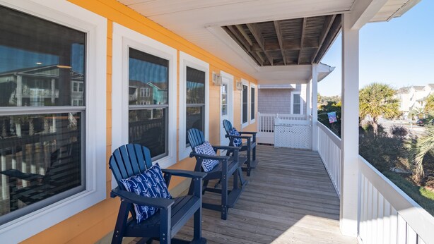 A covered porch with wooden flooring and white railings, featuring two Adirondack chairs with patterned cushions. - Legacy - A beautiful 4-bedroom home with a community dock, minutes from the ocean. (Holden Beach)
