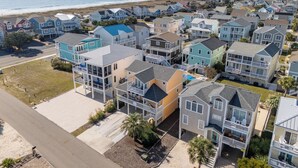 A coastal residential area with multi-story houses, a visible road, and a beach in the background. - Legacy - A beautiful 4-bedroom home with a community dock, minutes from the ocean. (Holden Beach)
