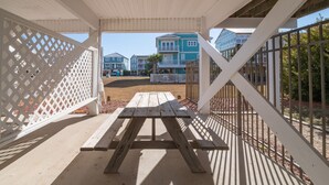 A covered outdoor area with a wooden picnic table, white lattice panels, and a view of buildings in the background. - Legacy - A beautiful 4-bedroom home with a community dock, minutes from the ocean. (Holden Beach)