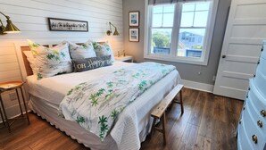 A bedroom with a bed, a bench, a dresser, and a window with a view of houses. - Hobbs Realty - Spend many an “Oh Happy Day” when you enjoy this charming home with a pool. (Holden Beach)