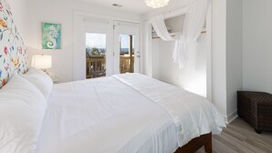 A bedroom with a large bed, a nightstand, a lamp, a window with a view, and a balcony. - See why this is “Our Happy Place” in this pet-friendly, oceanfront home with a hot tub. (Holden Beach)
