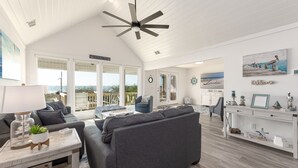A spacious living room with a large gray sofa, a wooden coffee table, and a ceiling fan. There is a view of the ocean through large windows. - See why this is “Our Happy Place” in this pet-friendly, oceanfront home with a hot tub. (Holden Beach)
