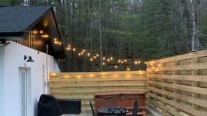 Une terrasse en bois avec un jacuzzi, une table et des chaises, entourée d’une clôture en bois et illuminée de guirlandes lumineuses.